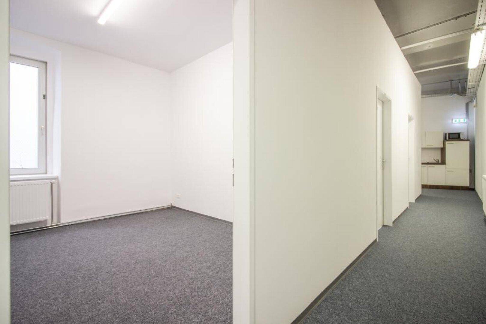 Büroraum mit grauem Teppichboden und Fenster, angrenzend an einen Flur mit weiteren Türen und einer Küchenzeile.