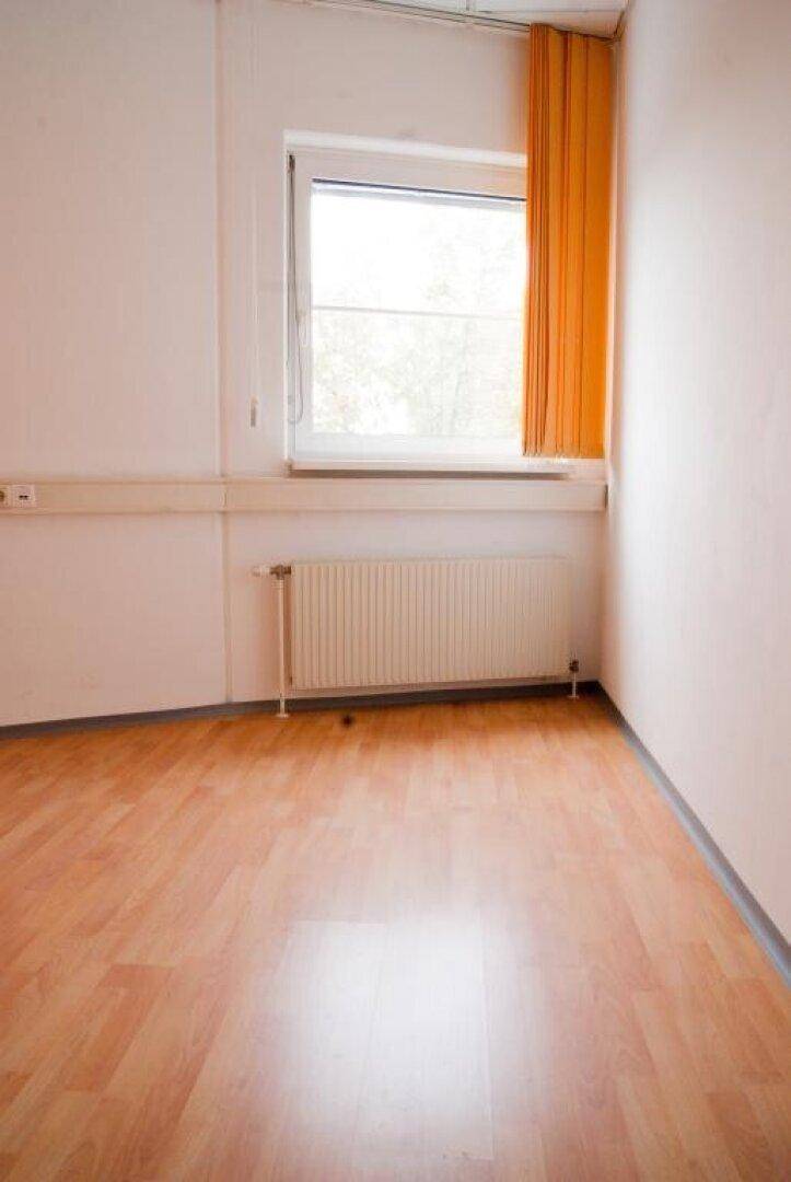 Ecke eines leeren Zimmers mit hellem Holzfußboden, weißer Wand und einem Fenster mit orangefarbenem Vorhang.