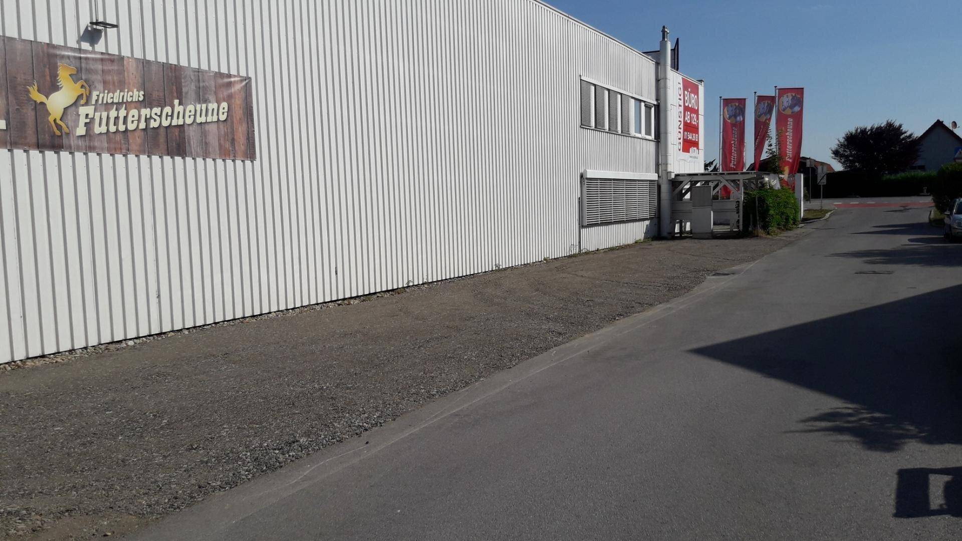 Seitenansicht eines weißen Gebäudes mit Wellblechfassade und einem Schild für 'Friedrichs Futterscheune' entlang einer Straße.
