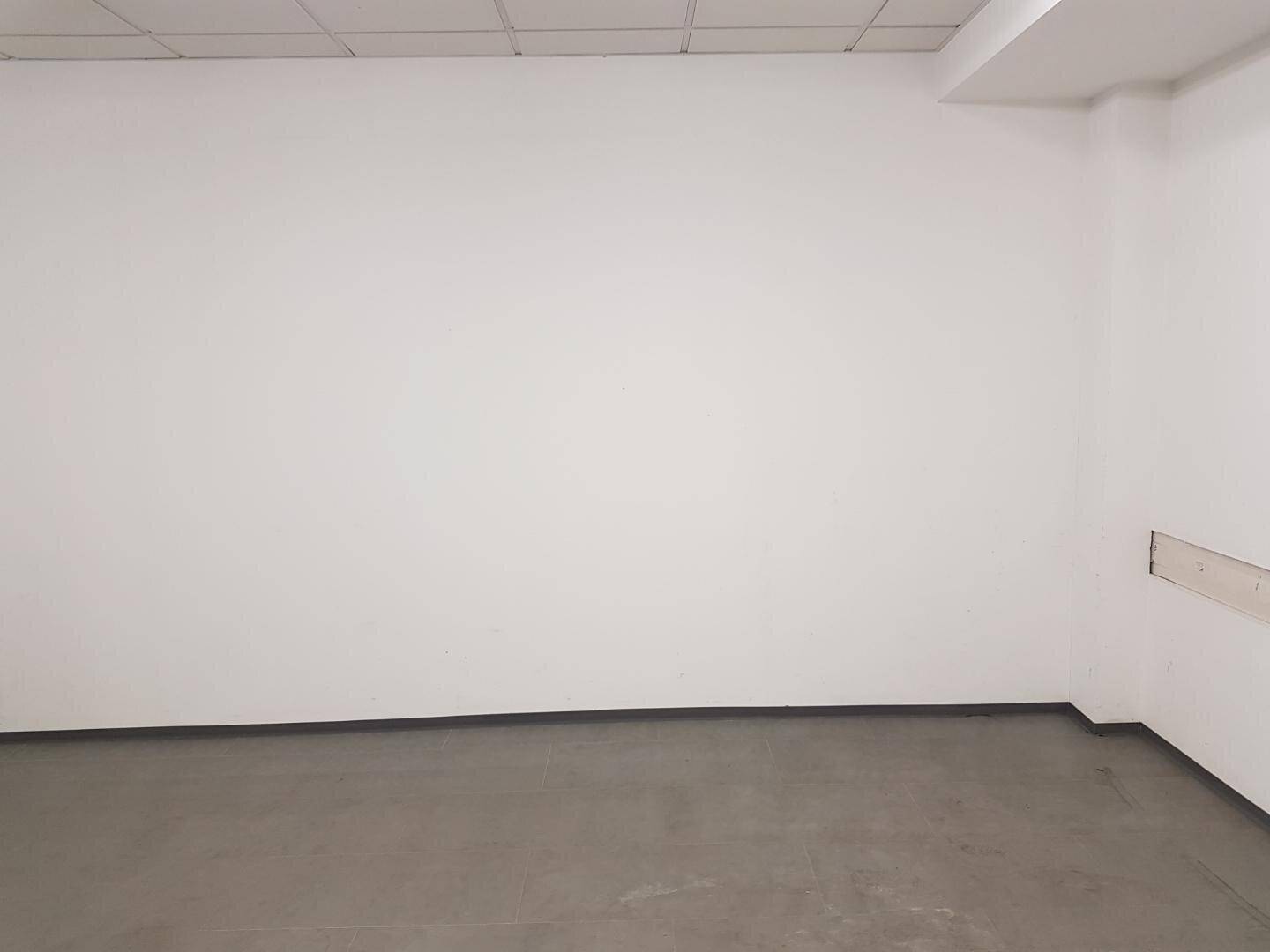 Leere Wand eines Gewerberaums mit weißem Anstrich und grauem Bodenbelag unter Deckenleuchten.