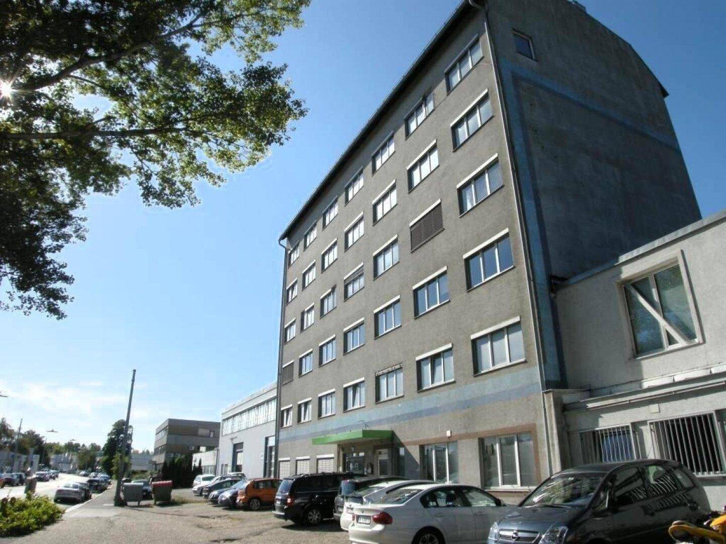 Modernes Bürogebäude mit mehreren Etagen, großen Fenstern und Parkplätzen davor unter blauem Himmel.