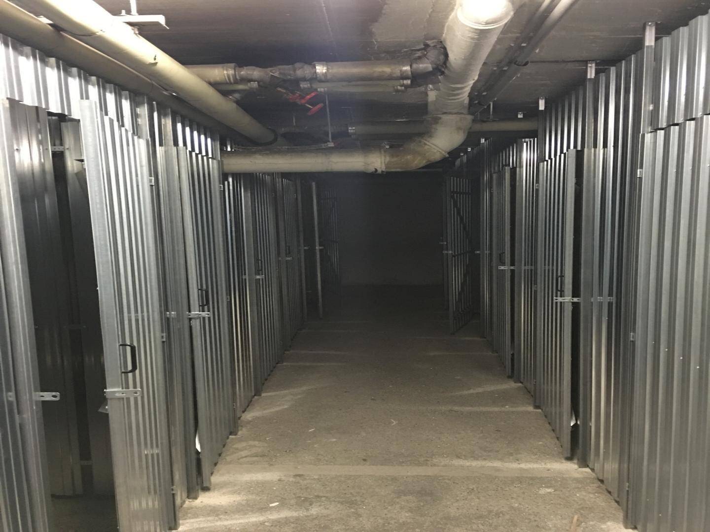 Langer Kellergang mit zahlreichen Metallabteilen für zusätzlichen Stauraum und sichtbaren Versorgungsleitungen.