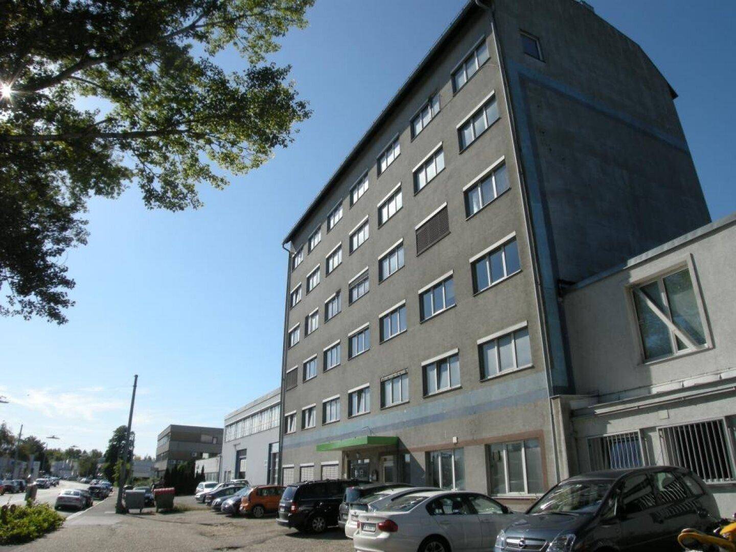 Mehrstöckiges Bürogebäude mit grauer Fassade, vielen Fenstern und einem Parkplatz davor unter blauem Himmel.