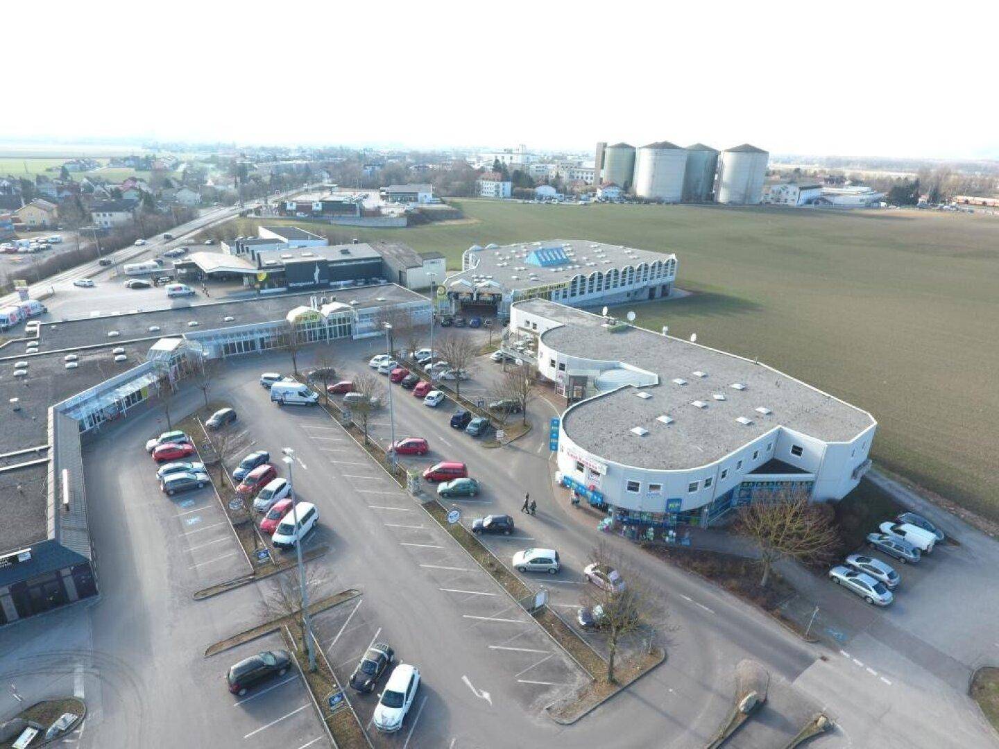 Luftaufnahme eines Gewerbegebiets mit mehreren Gebäuden, Parkplätzen und Industrieanlagen im Hintergrund.