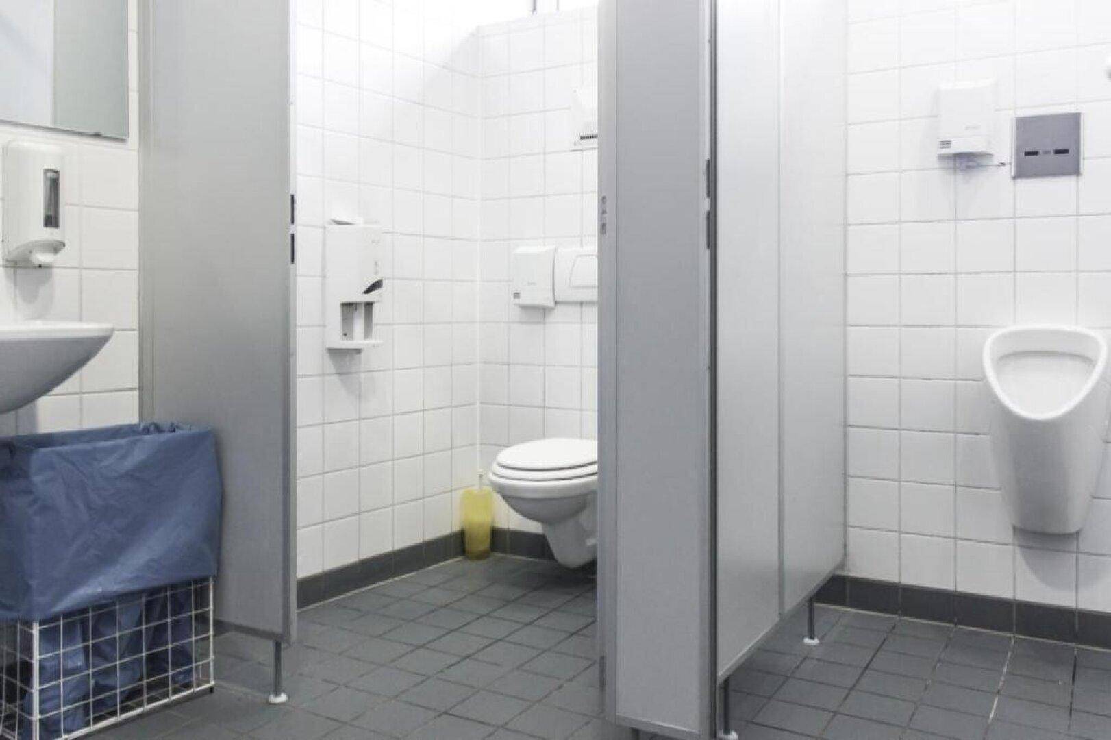 Öffentliche Toilette mit mehreren Kabinen, einem Urinal und einem Waschbecken, gefliest in Weiß und Grau.