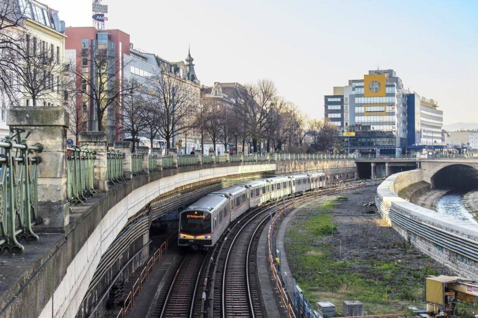 U-Bahn fährt durch eine städtische Landschaft mit Gebäuden und Brücken im Hintergrund.