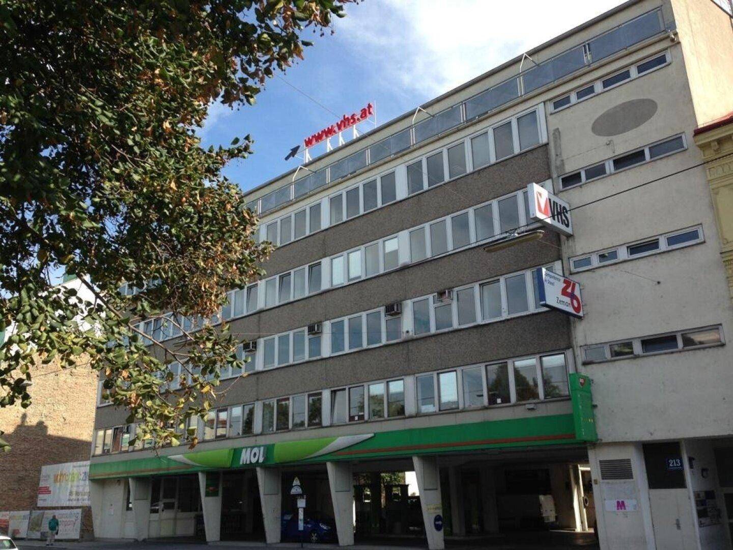 Mehrstöckiges Gebäude mit zahlreichen Fenstern und einer Mischfassade, umgeben von Bäumen unter blauem Himmel.