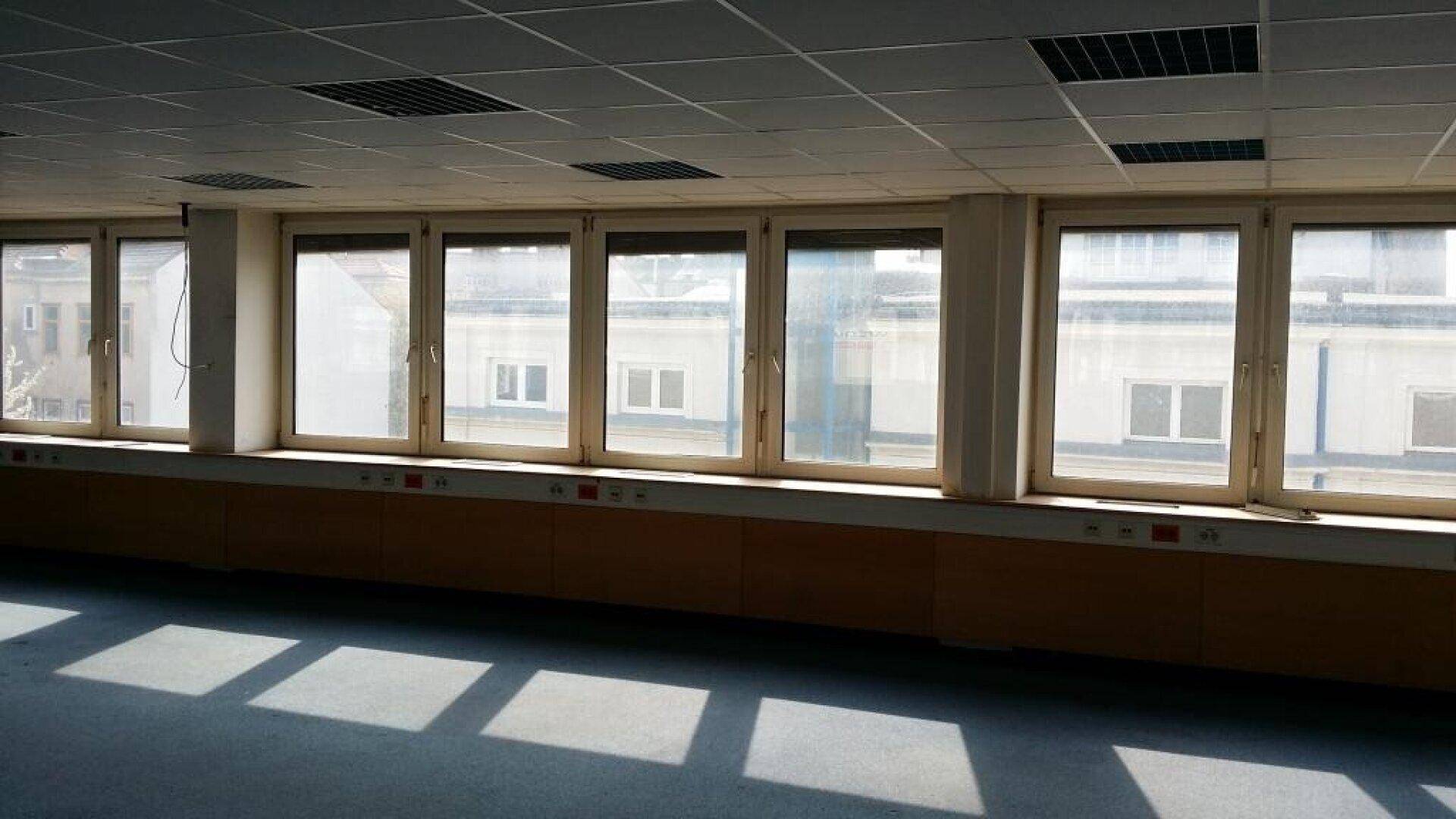 Großer, leerer Raum mit vielen Fenstern, die viel natürliches Licht hereinlassen, Teppichboden und Rasterdeckenplatten.