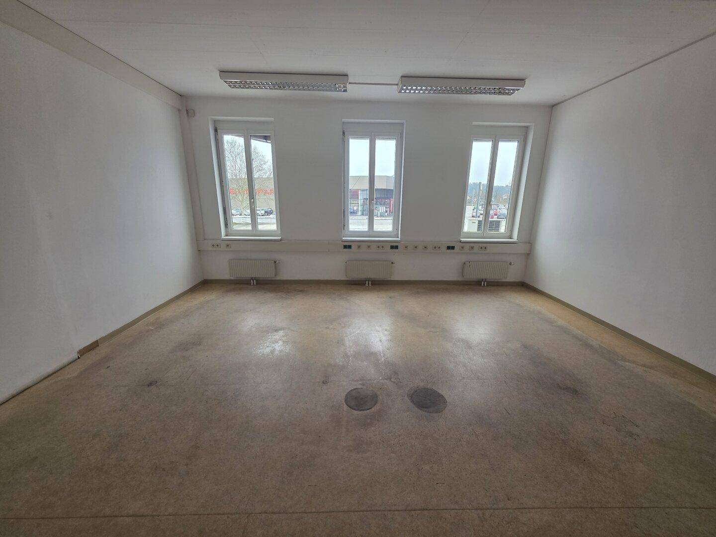 Heller, leerer Büroraum mit drei Fenstern und einfacher Ausstattung. Der Boden ist mit Vinyl belegt.