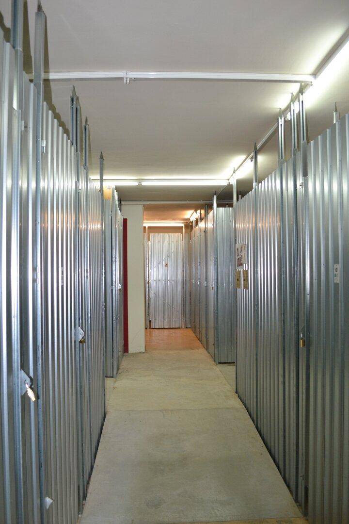 Langer Flur mit Reihen von Lagerabteilen und Deckenleuchten in einem Kellergeschoss.