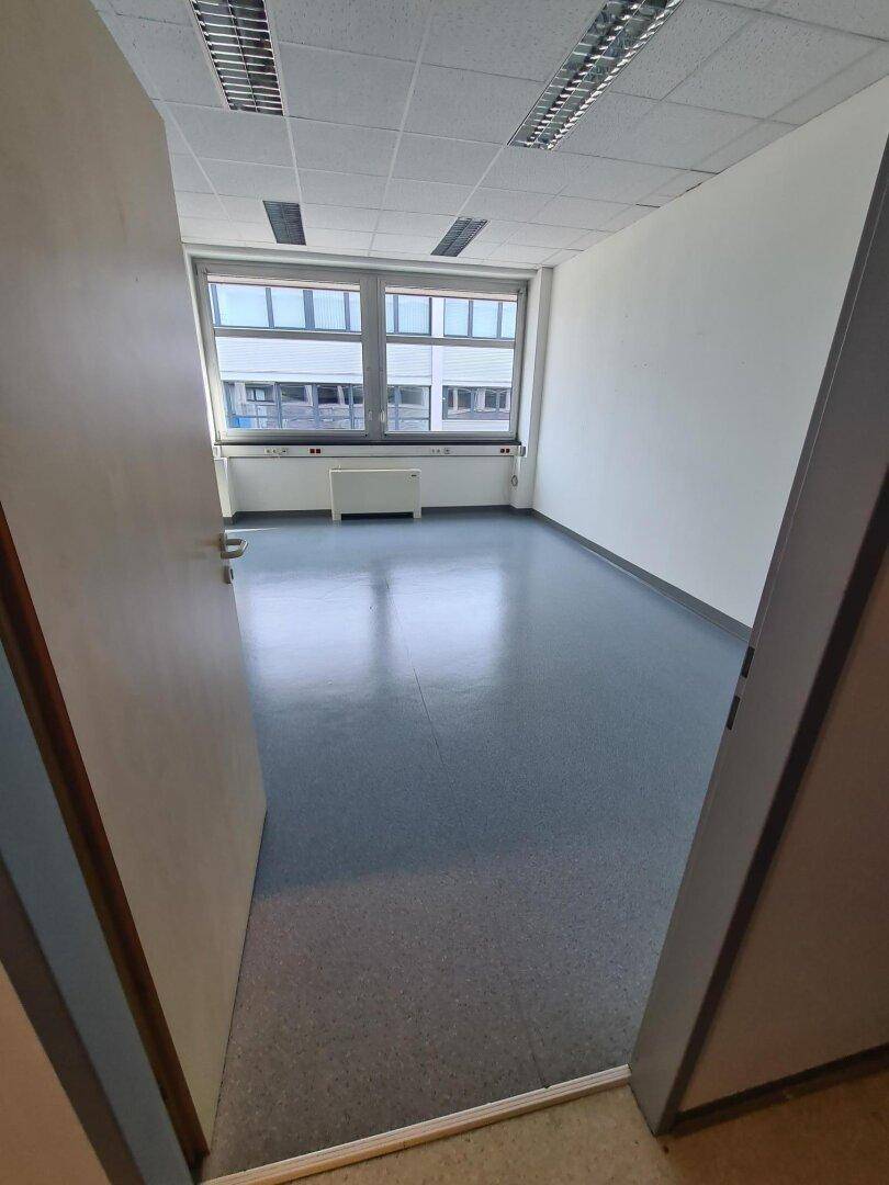 Leeres Büro mit grauen Vinylboden, großem Fenster und Heizkörper unter der Fensterbank.