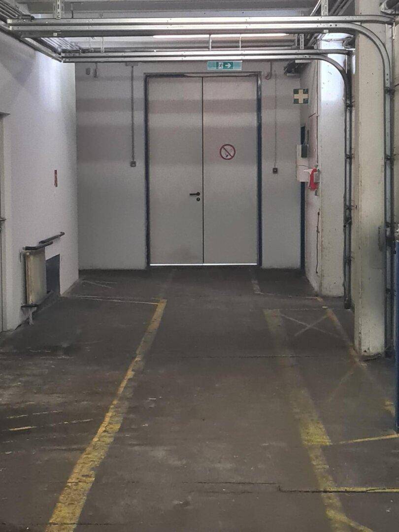 Industrieller Bereich mit großen Doppeltüren, sichtbaren Rohren und Betonboden.