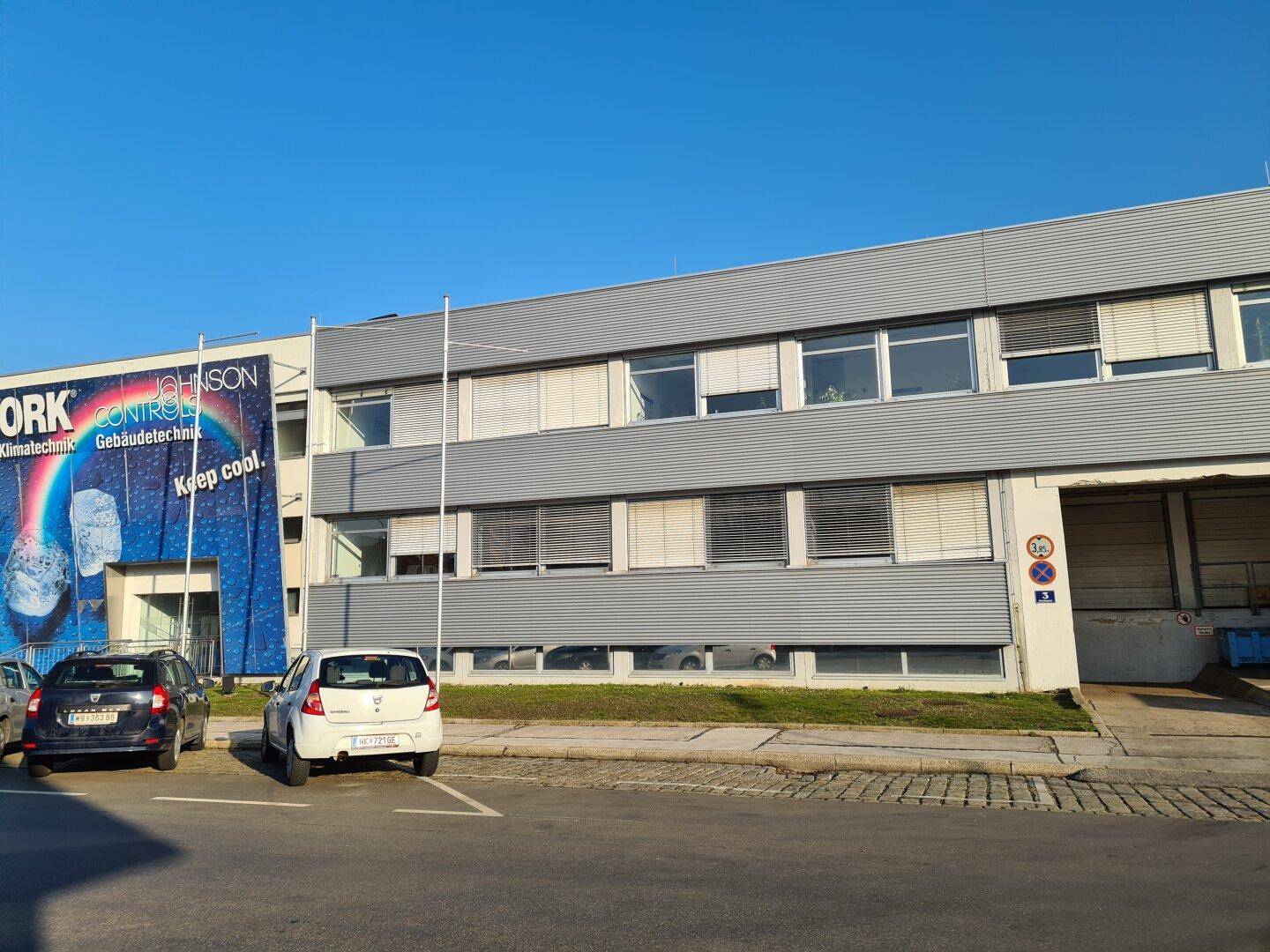 Gebäudeansicht mit Werbeplane, geparkten Autos und Fenstern mit Jalousien.