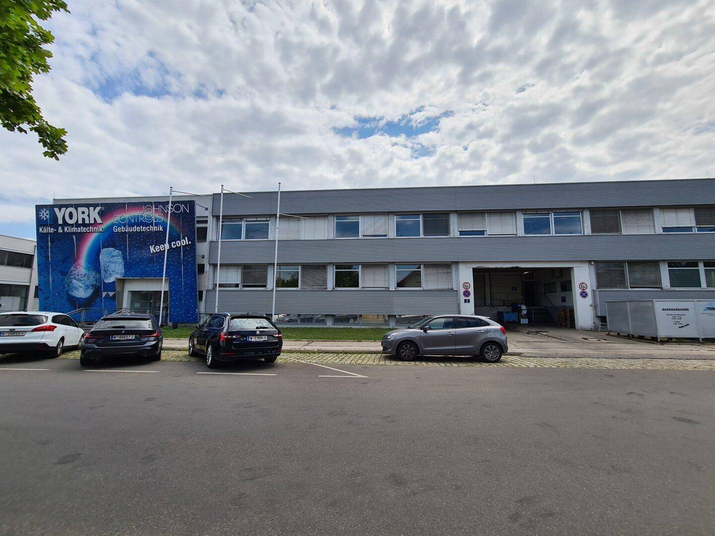 Außenansicht des Bürogebäudes mit Werbebanner und geparkten Fahrzeugen an einem bewölkten Tag.