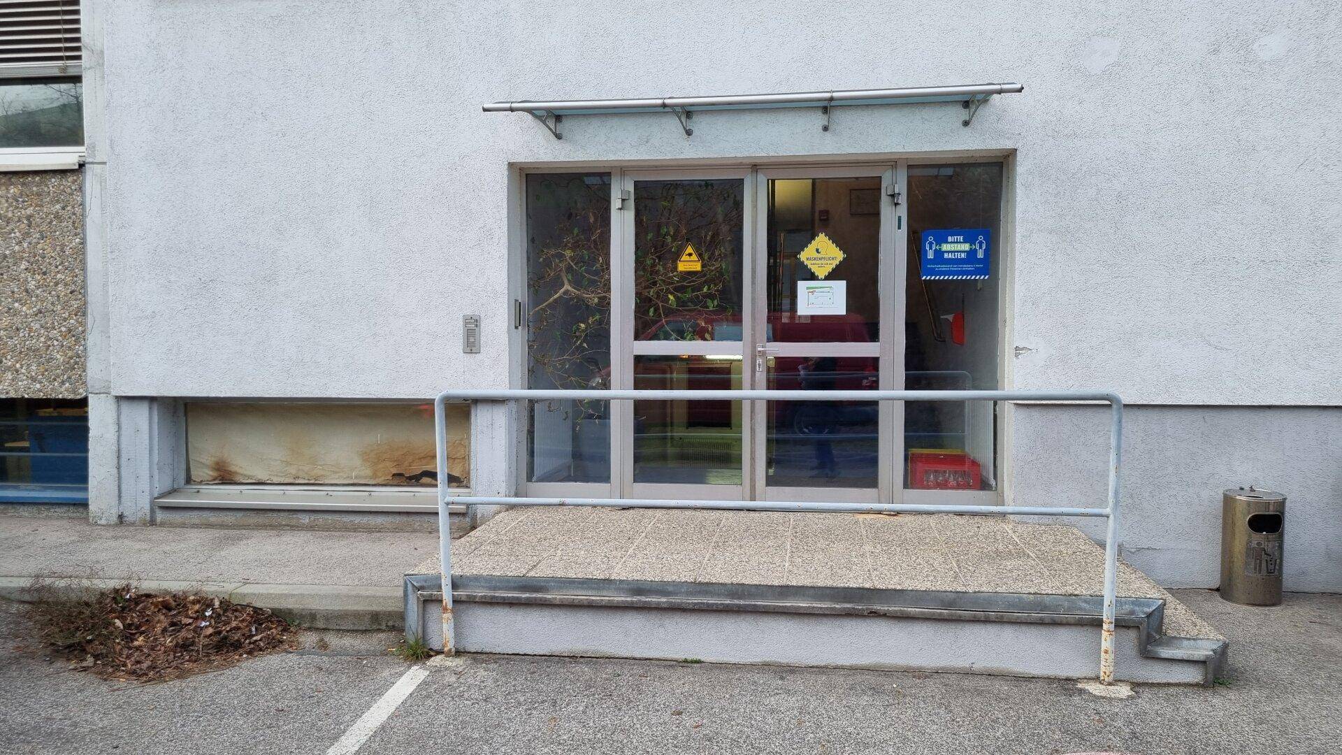 Nebeneingang des Gebäudes mit Glastüren, kleiner Rampe und einem Mülleimer.