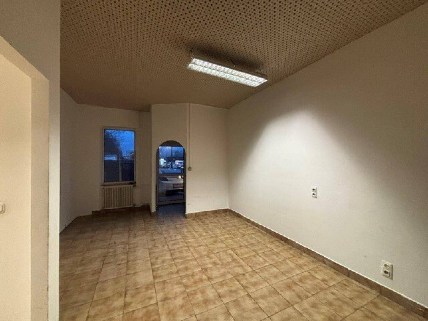 Leerer Raum mit braunen Fliesen, weißen Wänden, Fenster und perforierter Decke, mit Durchgang zu weiterem Bereich.