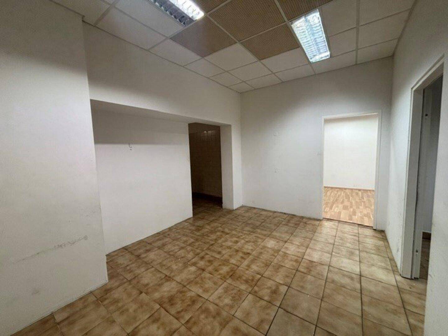 Geräumiger Raum mit braunen Fliesen, weißen Wänden und abgehängter Decke, mit Zugängen zu mehreren Zimmern.