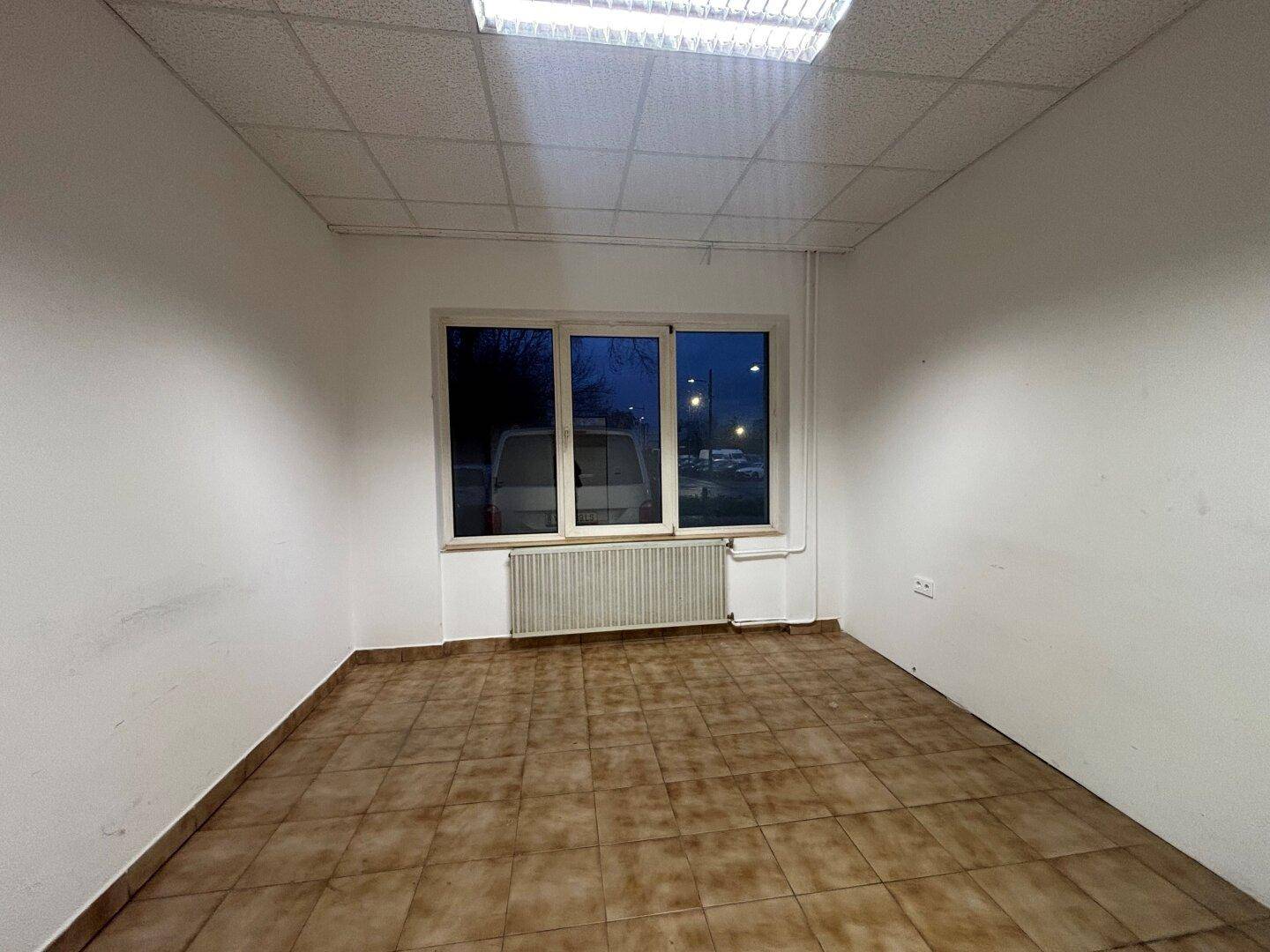 Heller Raum mit braunen Fliesen, weißen Wänden, großem Fenster und abgehängter Decke mit Leuchtstofflampen.
