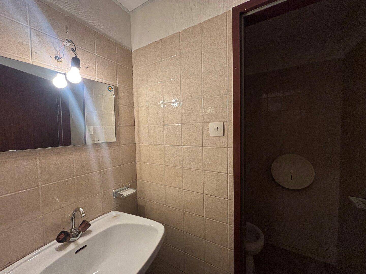 Badezimmerbereich mit weißem Waschbecken, Spiegel und beigefarbenen Fliesen an den Wänden, mit Blick auf eine Toilette.