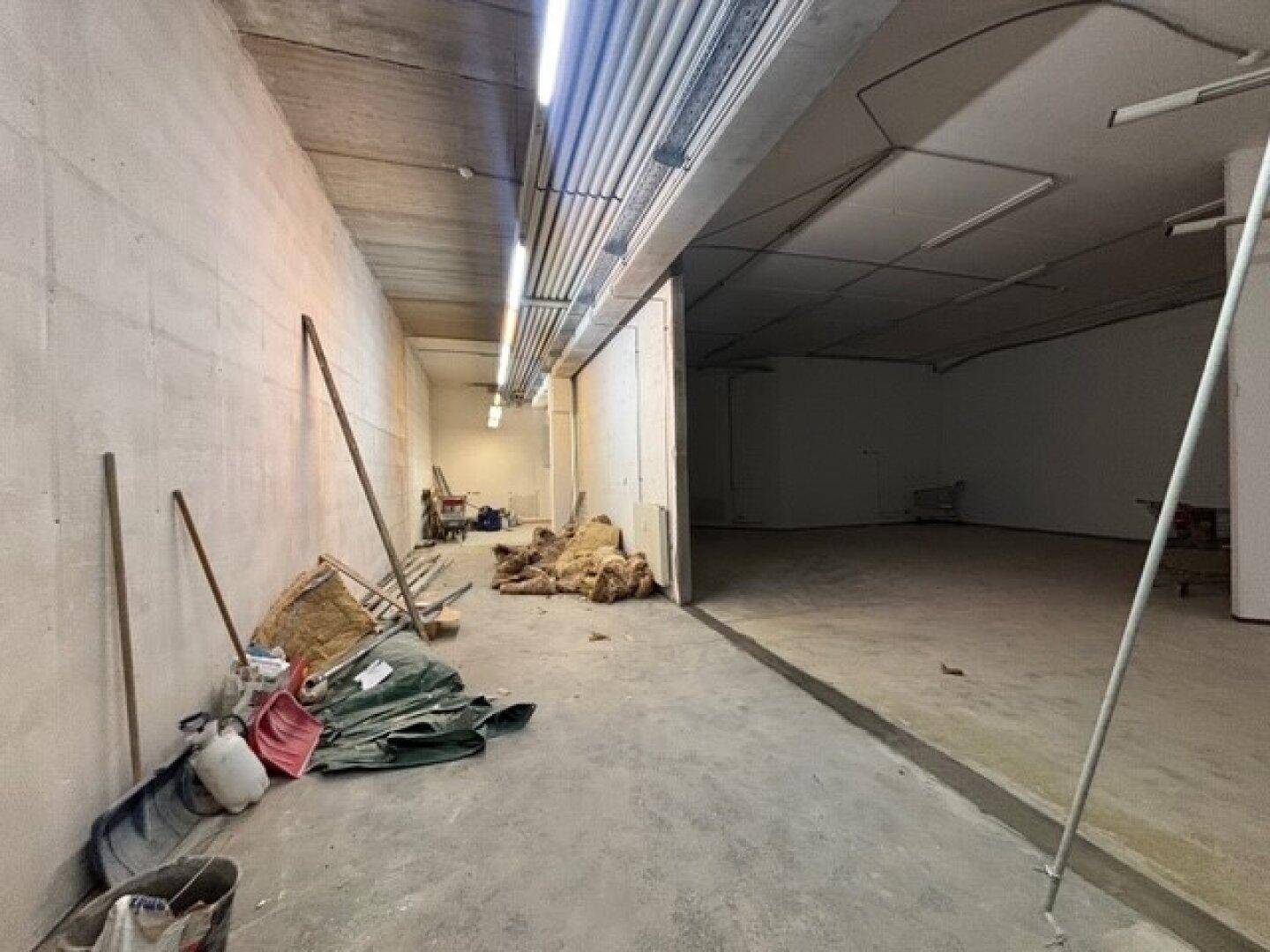 Perspektive eines langen, leeren Raumes mit Bauabfällen und sichtbaren Deckeninstallationen, die auf Fertigstellung warten.