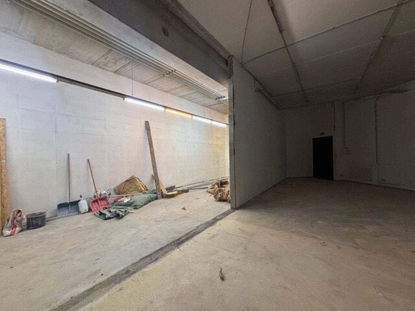 Langer, offener Innenbereich mit Betonboden, sichtbaren Deckenleuchten und Baumaterialien, die auf eine Renovierung warten.