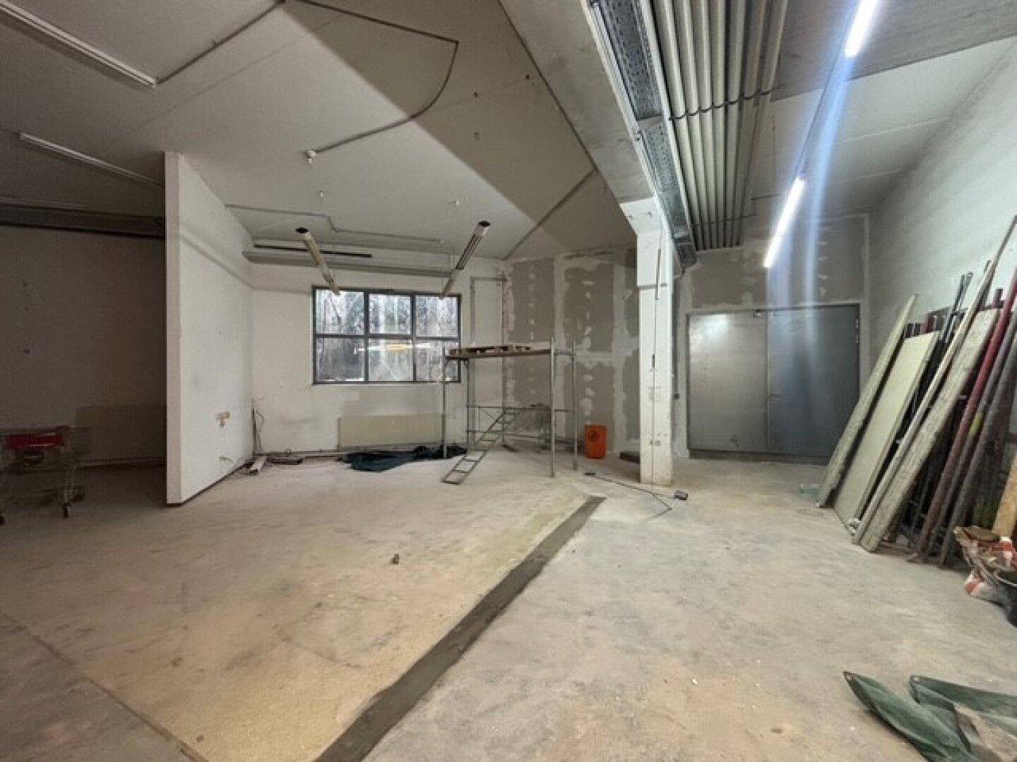 Weiterer Blick in den großen Innenraum mit Baugerüst, sichtbaren Leitungen und Fenstern, die Tageslicht hereinlassen.