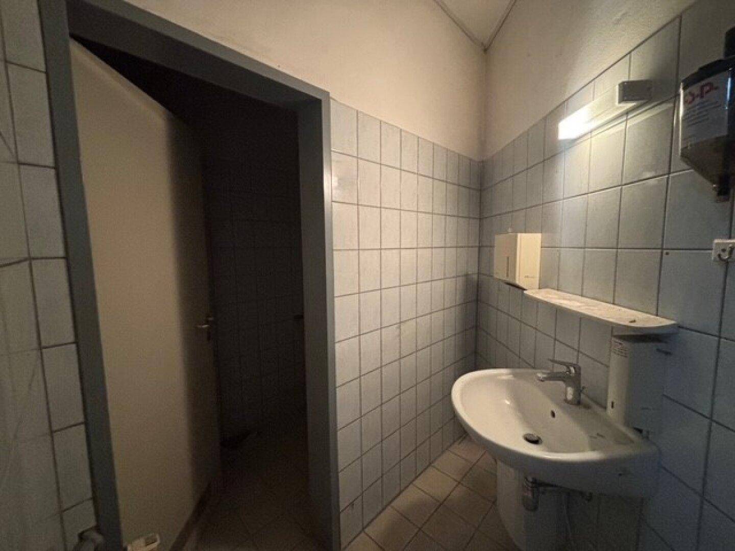 Einfaches Badezimmer mit hellblauen Fliesen, Waschbecken und einer Tür zum Nebenraum, das eine Modernisierung benötigt.