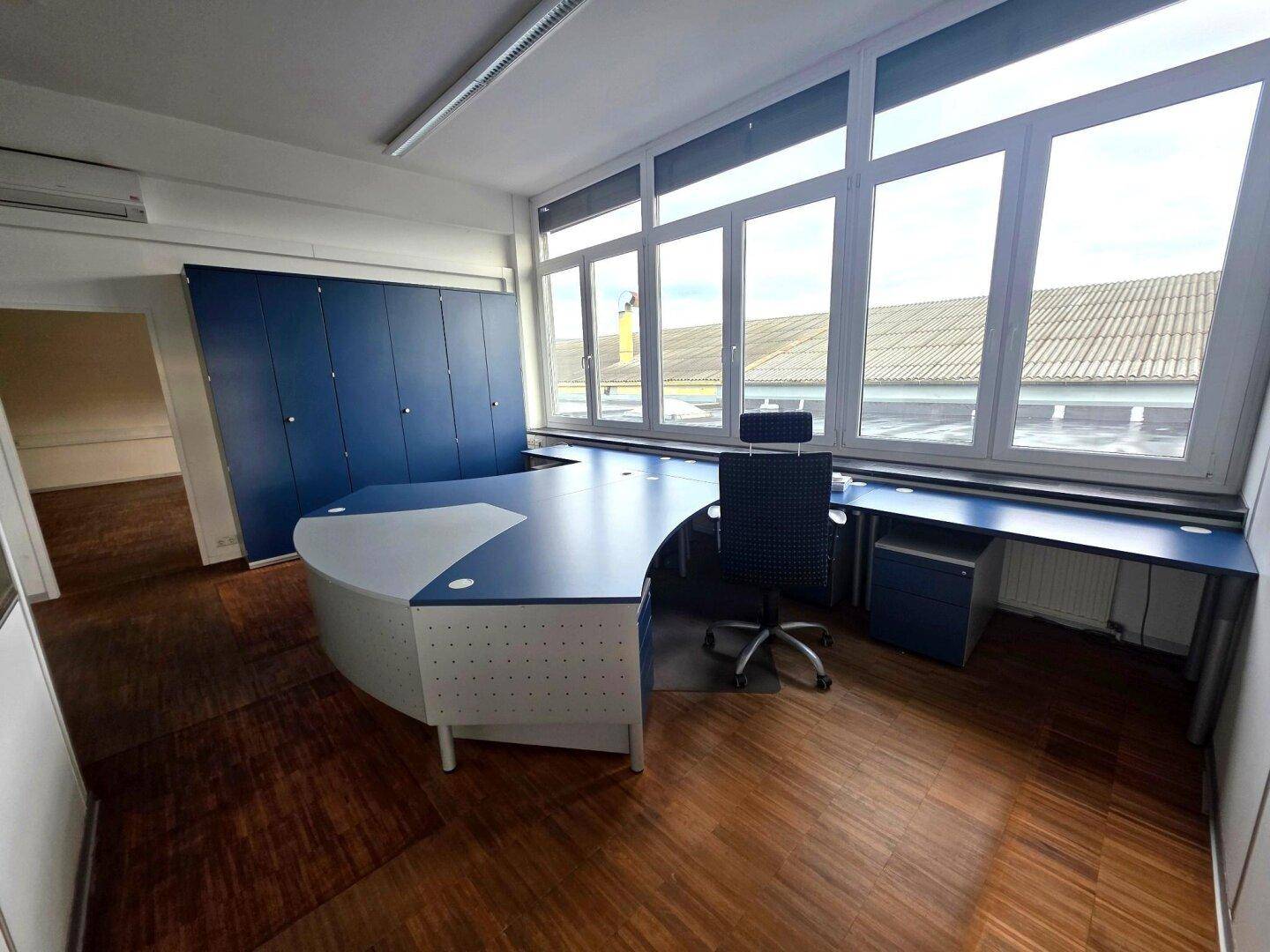 Modern eingerichtetes Büro mit blauem Schreibtisch, passenden Schränken und großen Fenstern.