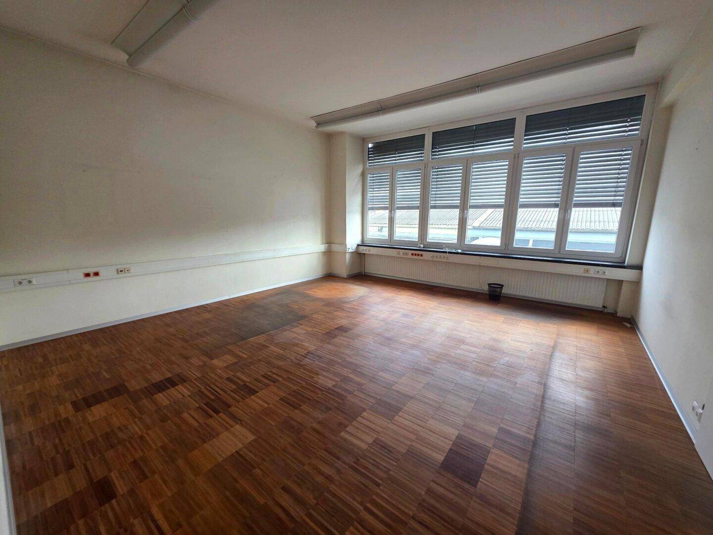 Leerer Büroraum mit Holzfußboden, großen Fenstern und weißen Wänden, bereit zur individuellen Gestaltung.