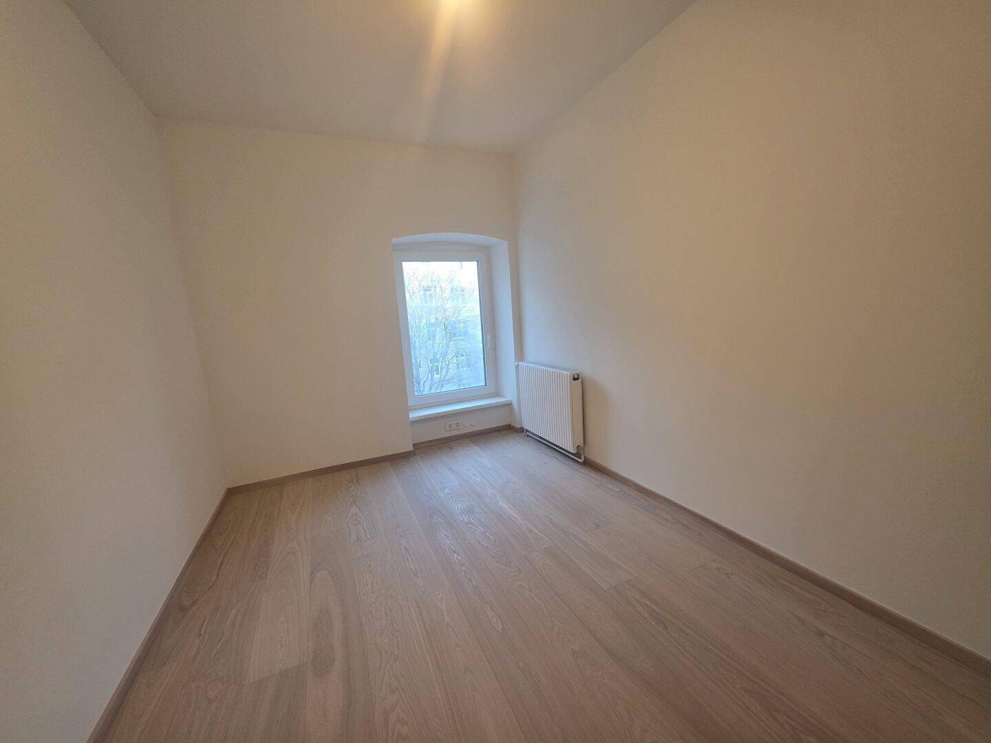 Gemütliches Schlafzimmer mit hellem Holzparkett, weiß gestrichenen Wänden und einem Fenster.