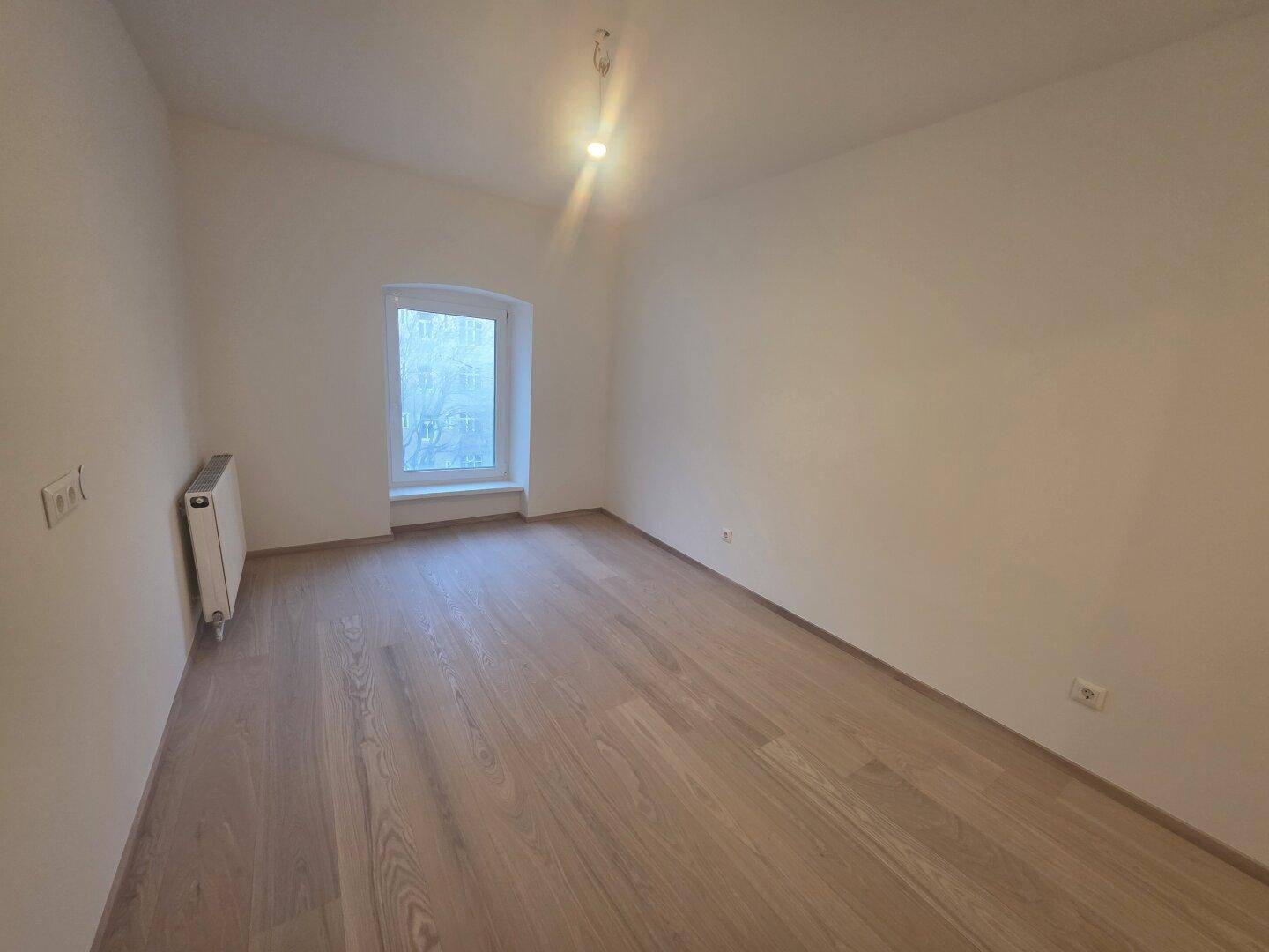 Helles Zimmer mit neuem Holzparkettboden, weißen Wänden und einem Fenster mit Heizkörper.