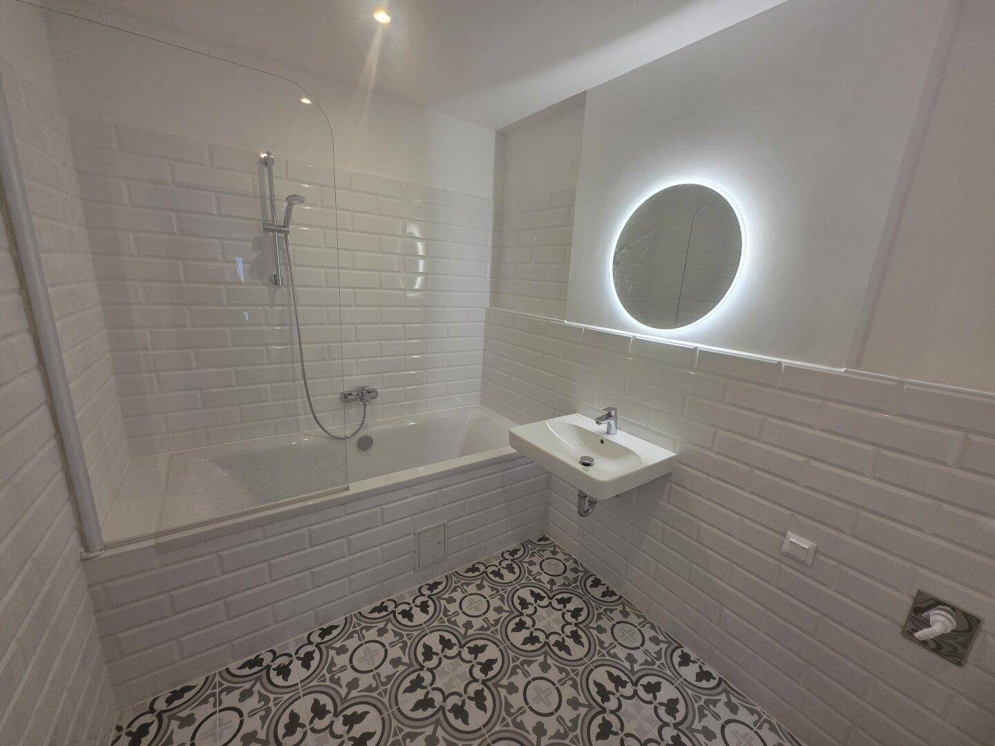 Modernes Badezimmer mit weißen Metrofliesen, gemustertem Boden und beleuchtetem Spiegel über dem Waschbecken.