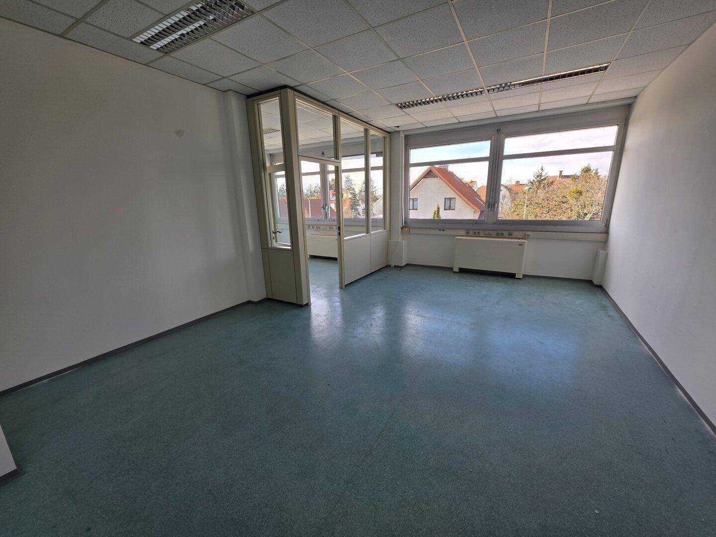 Bürofläche mit großen Fenstern und Blick auf Wohngebäude, teilweise durch Glaswände abgetrennt.