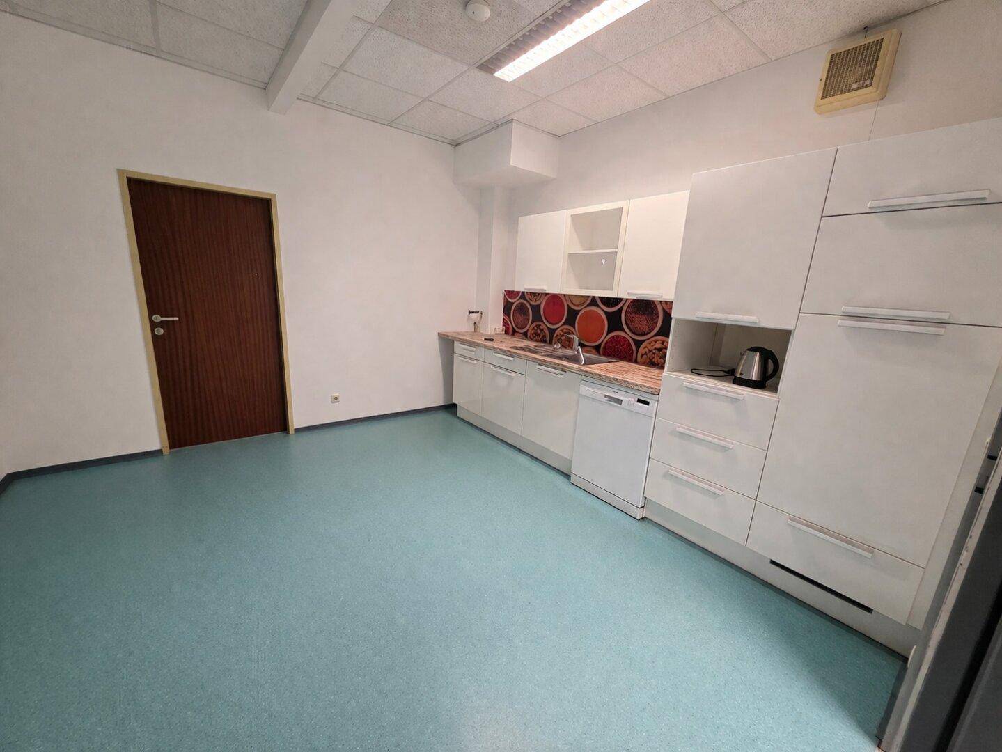 Funktionale Küchenzeile mit weißen Schränken, Spülmaschine und farbiger Spritzschutzwand in einem Büroraum.