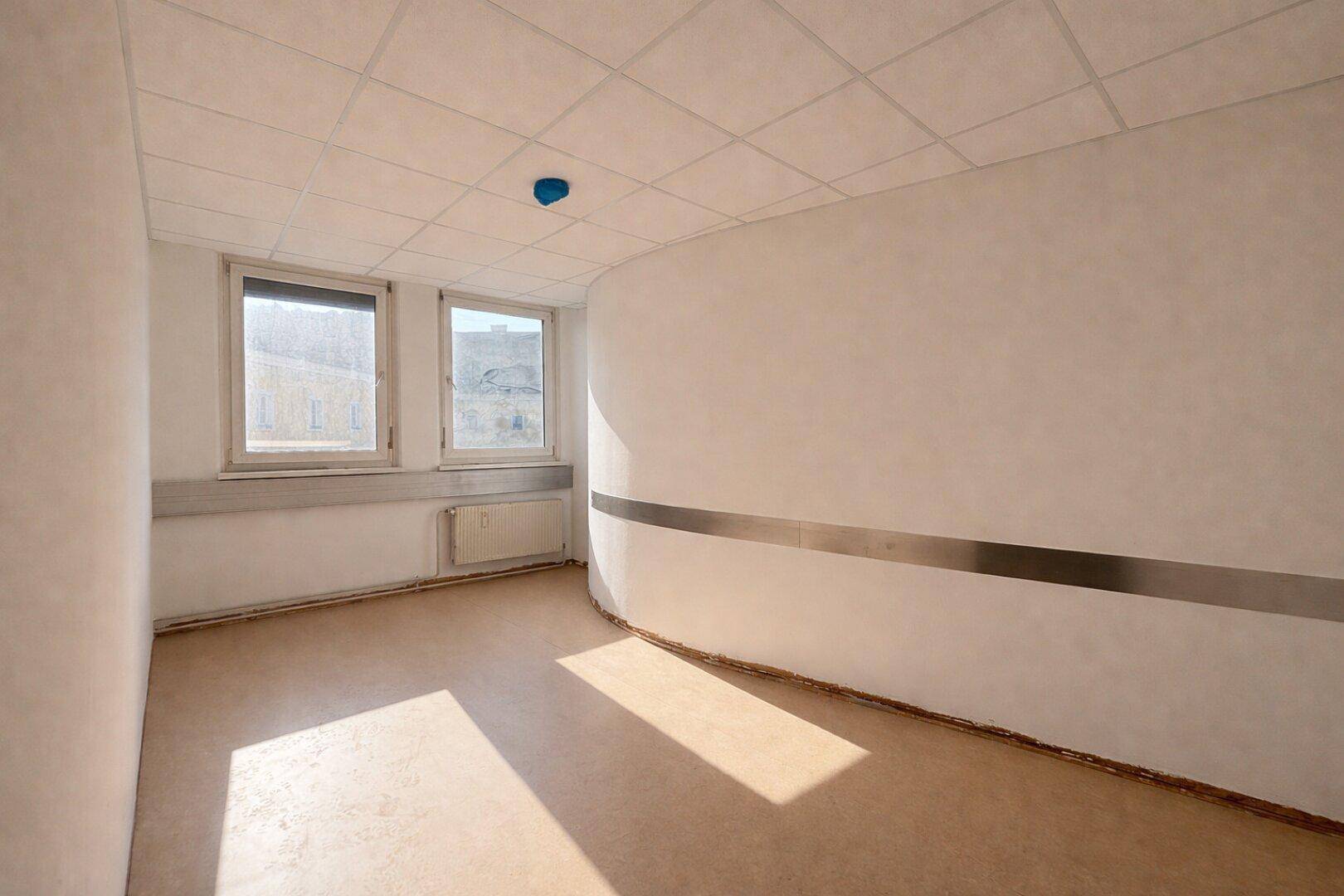 Leerer Büroraum mit zwei Fenstern, einer geschwungenen Wand und hellem Bodenbelag, gut beleuchtet.