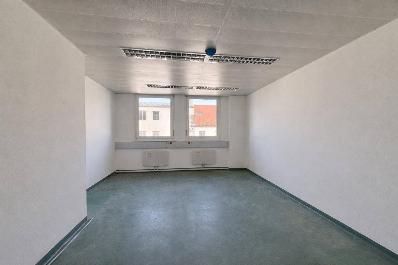 Leerer Büroraum mit zwei Fenstern, weiß gestrichenen Wänden und dunklem, pflegeleichtem Bodenbelag.