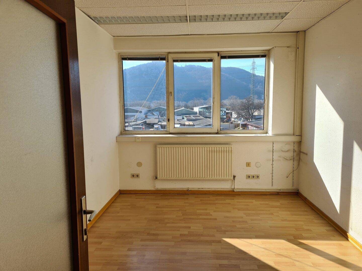 Leerer Büroraum mit Laminatboden, Heizkörper und einem weiten Blick auf Berge und Umgebung.