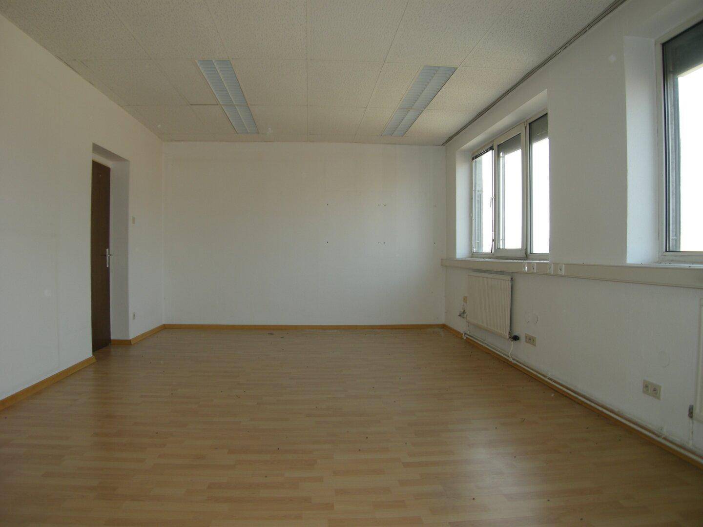 Geräumiger, leerer Büroraum mit Laminatboden, weißen Wänden und Deckenplatten, bereit zur Nutzung.