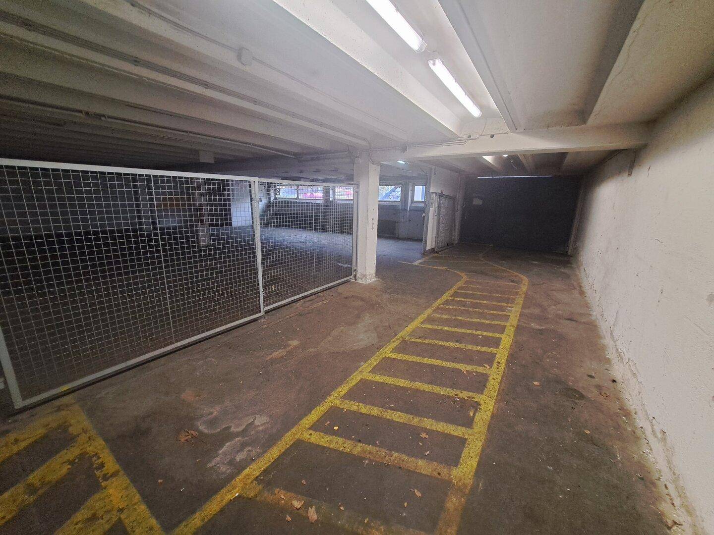 Tiefgarage mit gelben Bodenmarkierungen und Gitterabtrennung, beleuchtet von Deckenleuchten.