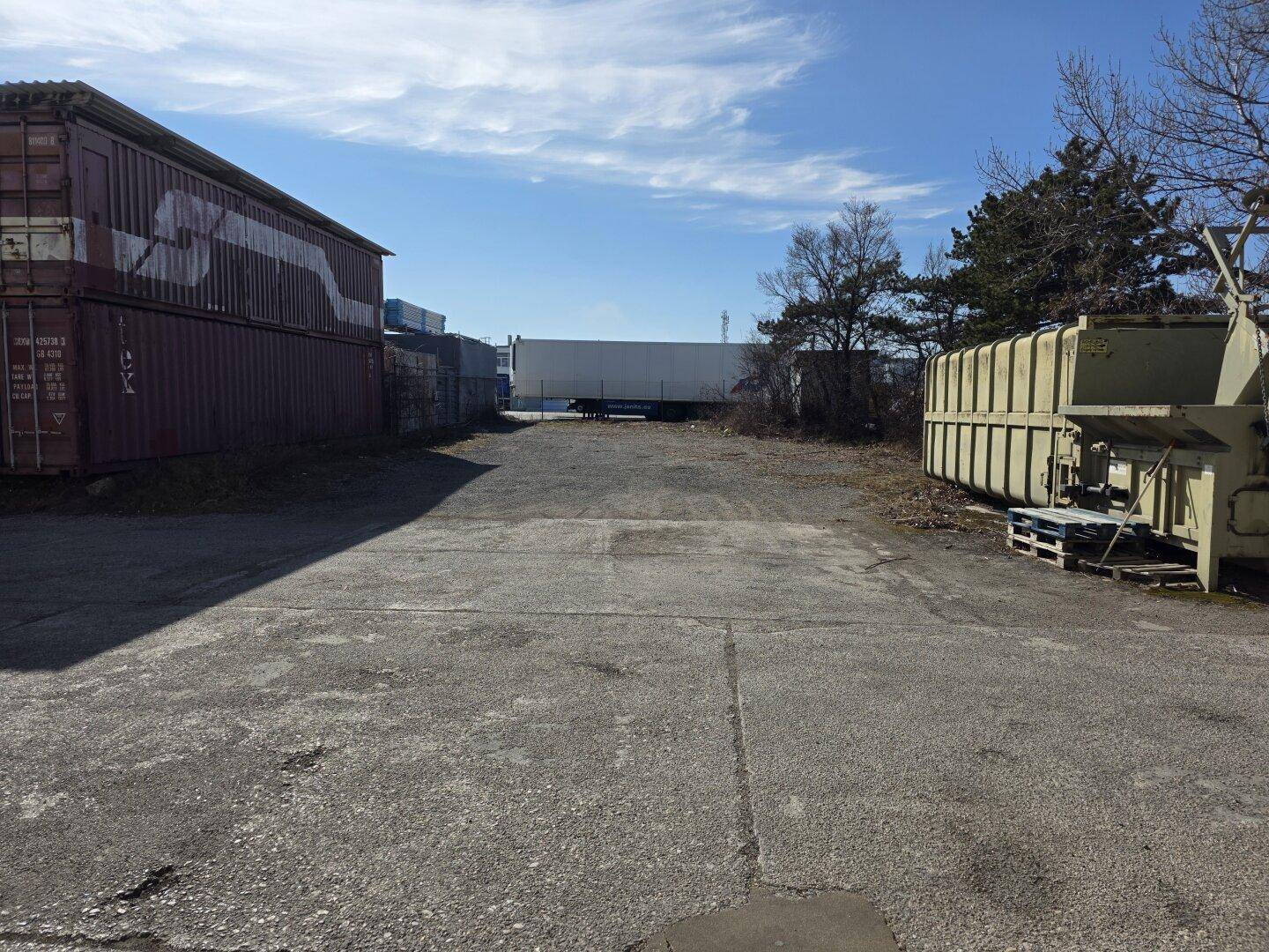 Außenbereich eines Industriegeländes mit Lagercontainern und einem großen Müllcontainer unter blauem Himmel.