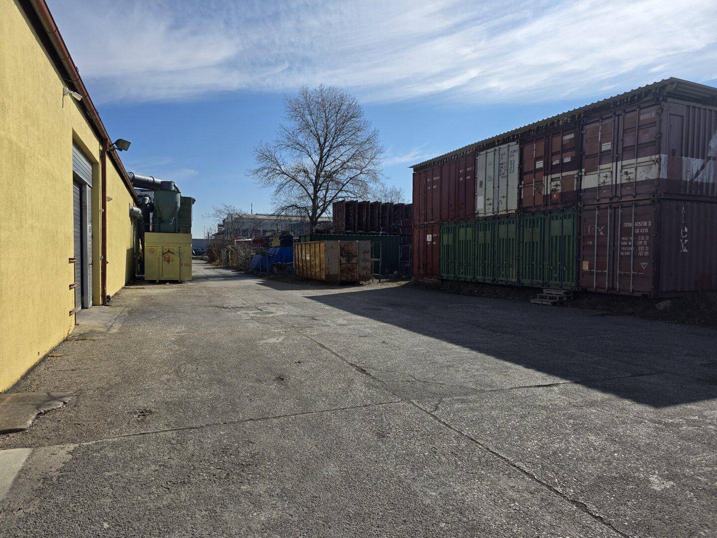Blick auf ein Industriegelände mit Lagercontainern, einem gelben Gebäude und einer asphaltierten Zufahrt.