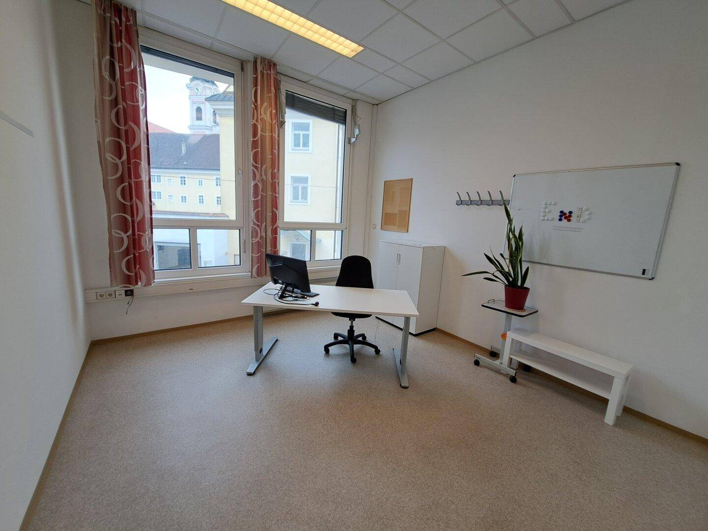 Helles Büro mit großen Fenstern, Blick auf ein historisches Gebäude und einfacher, funktionaler Einrichtung.