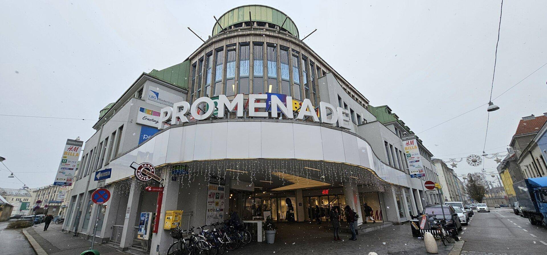 Außenansicht eines modernen Geschäftsgebäudes namens 'PROMENADE' an einer belebten Straße im Winter.