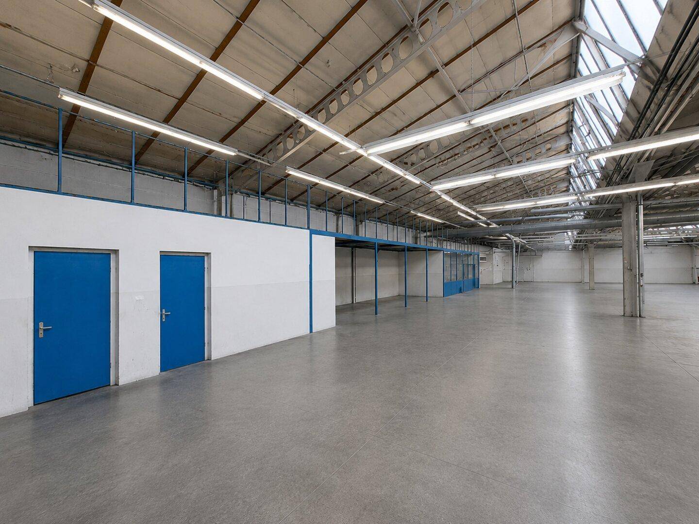 Geräumige Lagerhalle mit Betonboden, hohen Decken, blauen Türen und einer Zwischenebene.