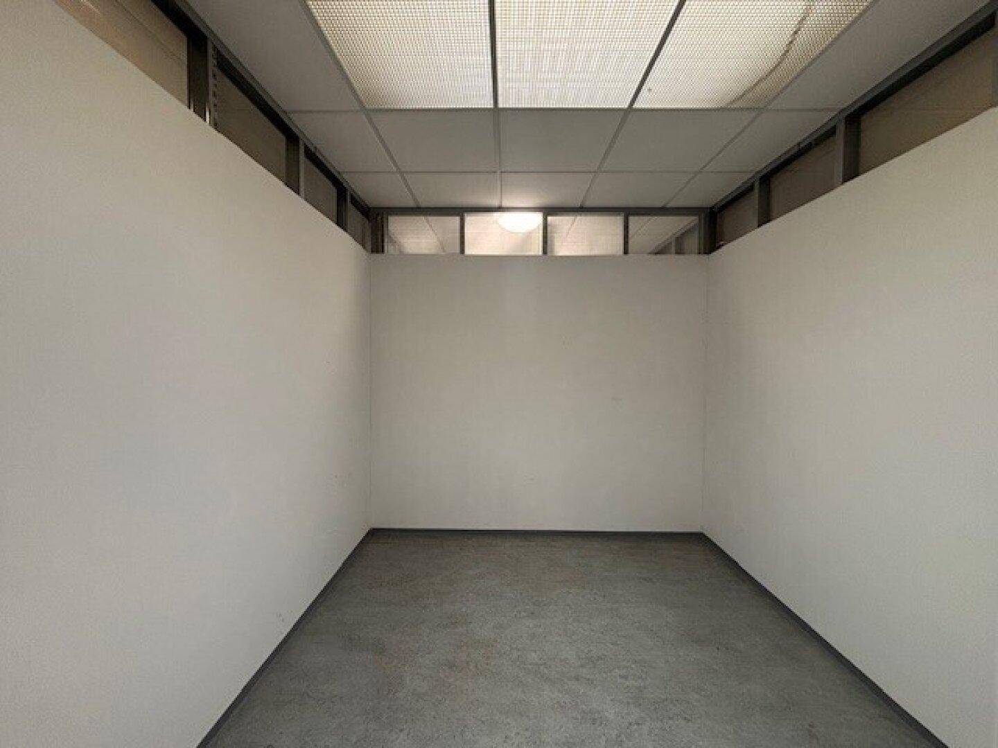 Leerer Raum mit weißen Wänden, grauem Betonboden und einer Rasterdecke mit Oberlichtern.