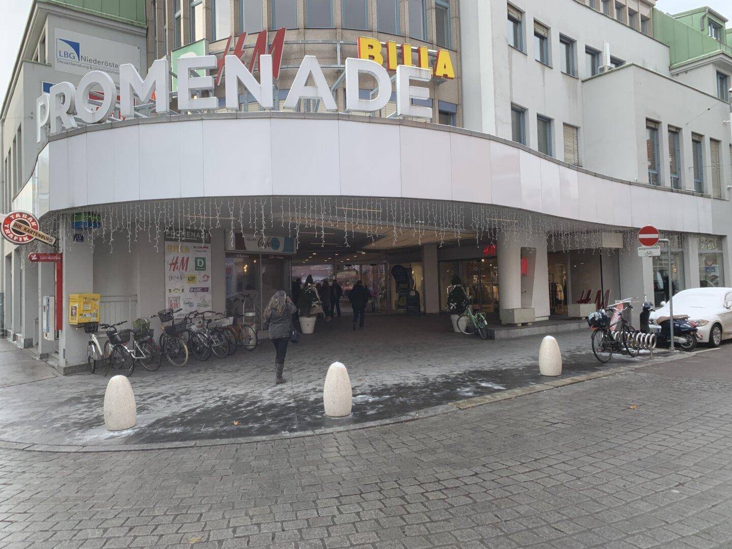 Eingangsbereich eines Einkaufszentrums von der Straße aus gesehen, mit Weihnachtsbeleuchtung und Passanten.