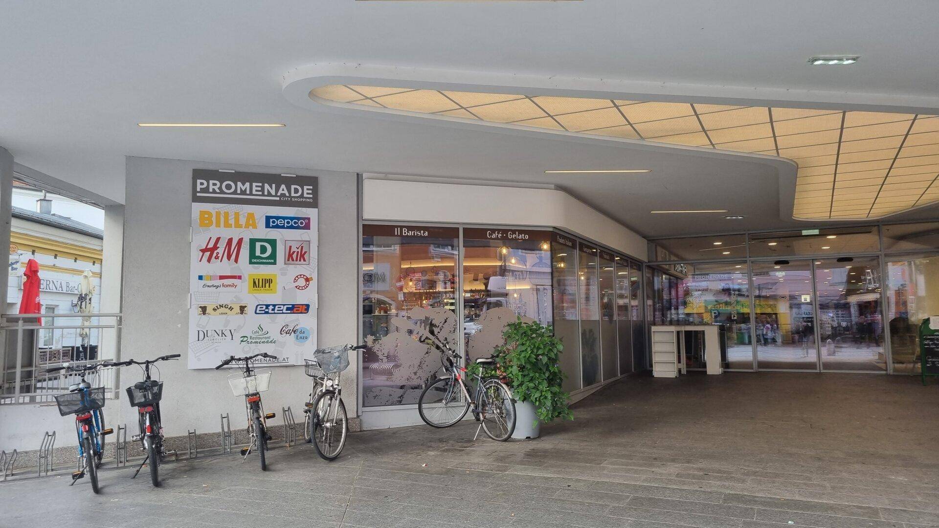Eingangsbereich eines Einkaufszentrums mit Fahrradständern und einer Übersichtstafel der Geschäfte.