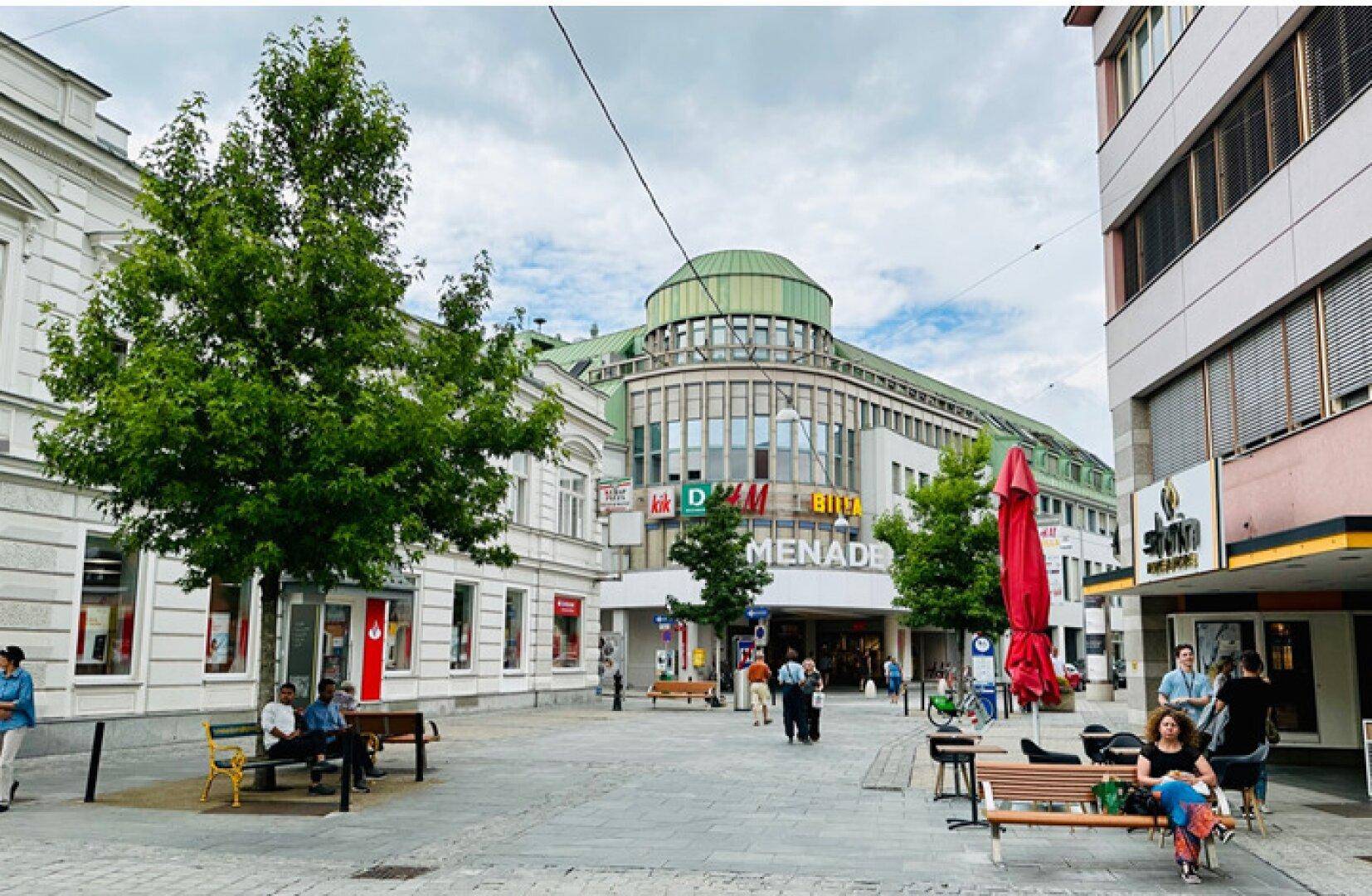 Stadtplatz mit Bäumen und Bänken vor dem Einkaufszentrum, belebt mit Passanten an einem Sommertag.