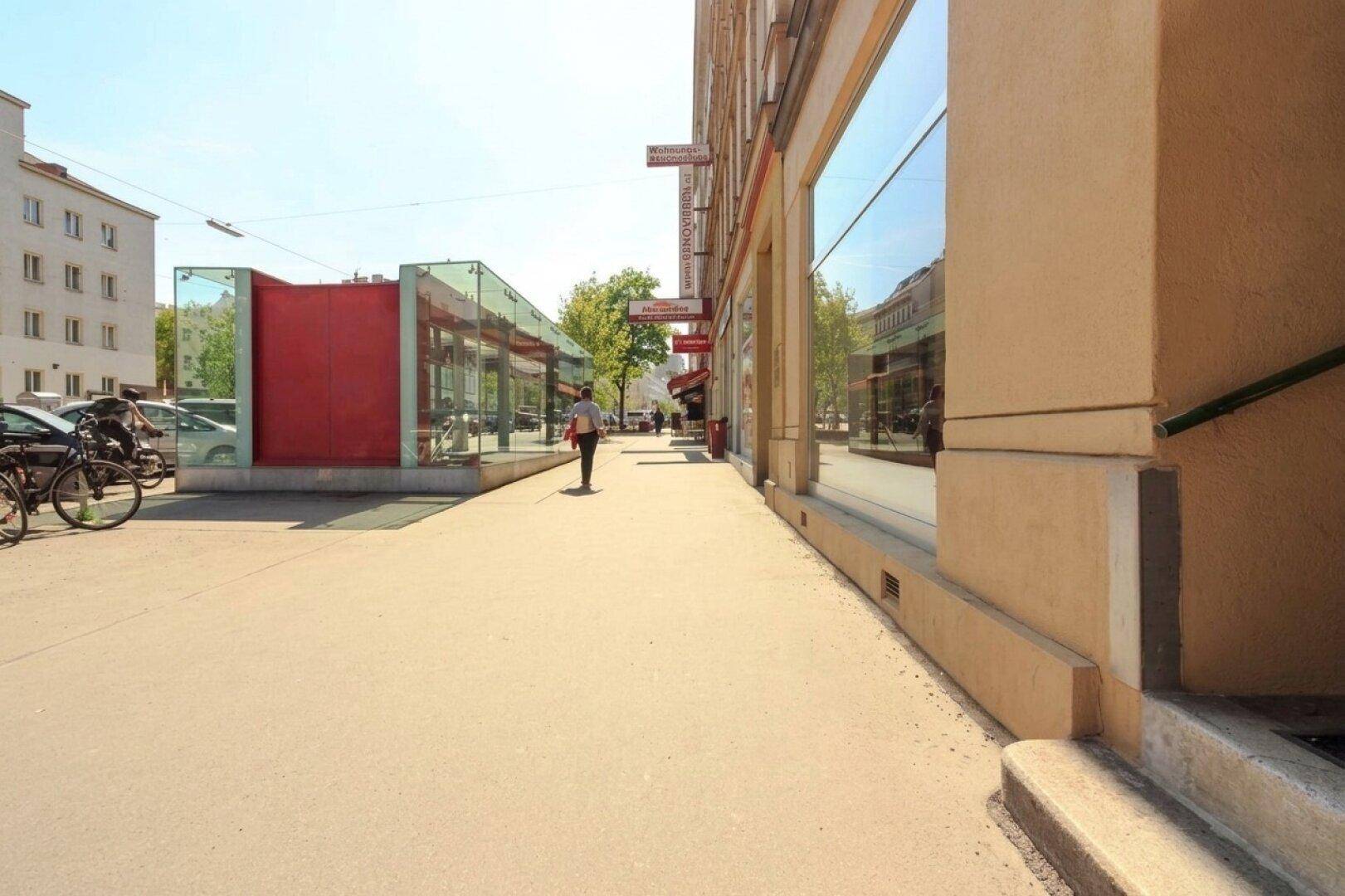 Blick auf eine belebte Straße mit einem Gebäude, großen Schaufenstern und einer modernen Glasstruktur an einem sonnigen Tag.