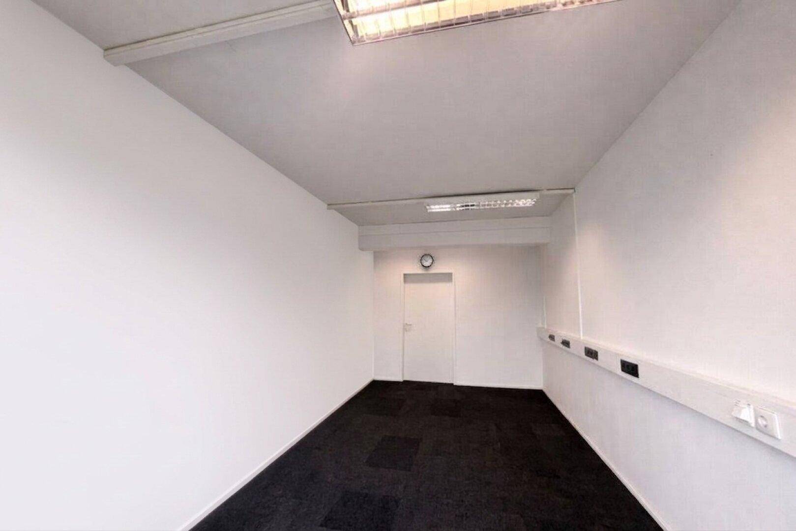 Leerer Büroraum mit weißen Wänden, dunklem Teppichboden und Deckenbeleuchtung sowie einem Oberlicht.
